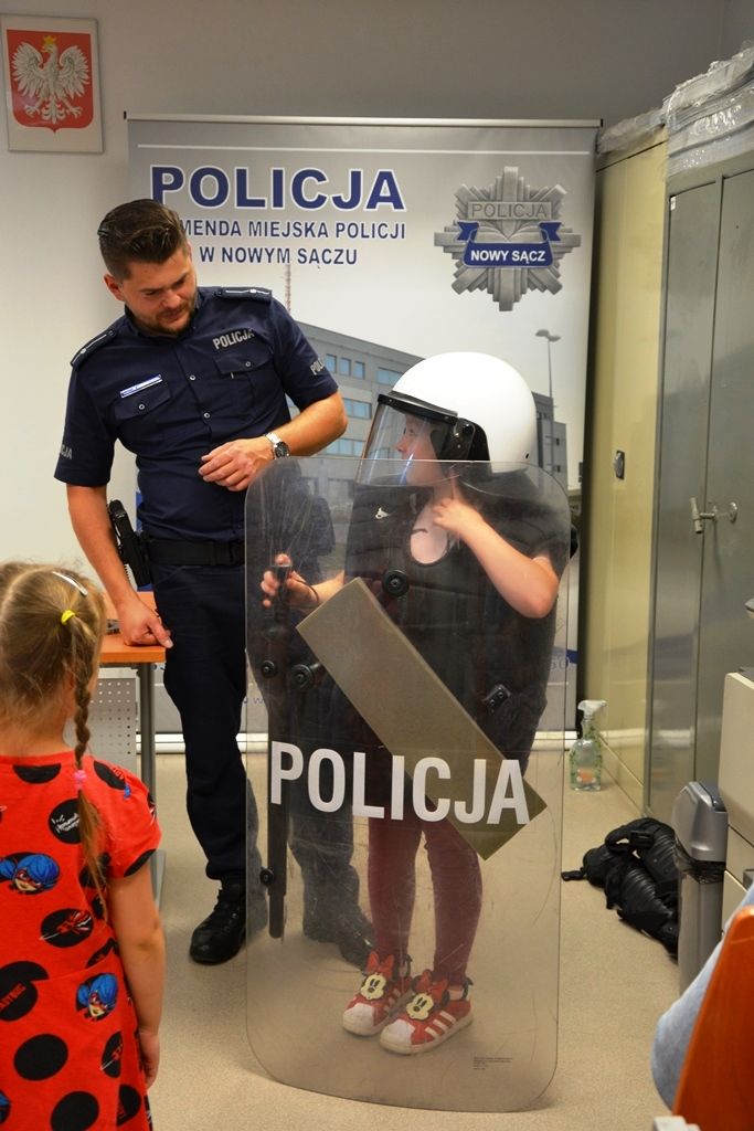 dziewczynka w policyjnym kasku ochronnym z tarczą w ręku, obok policjant prewencji