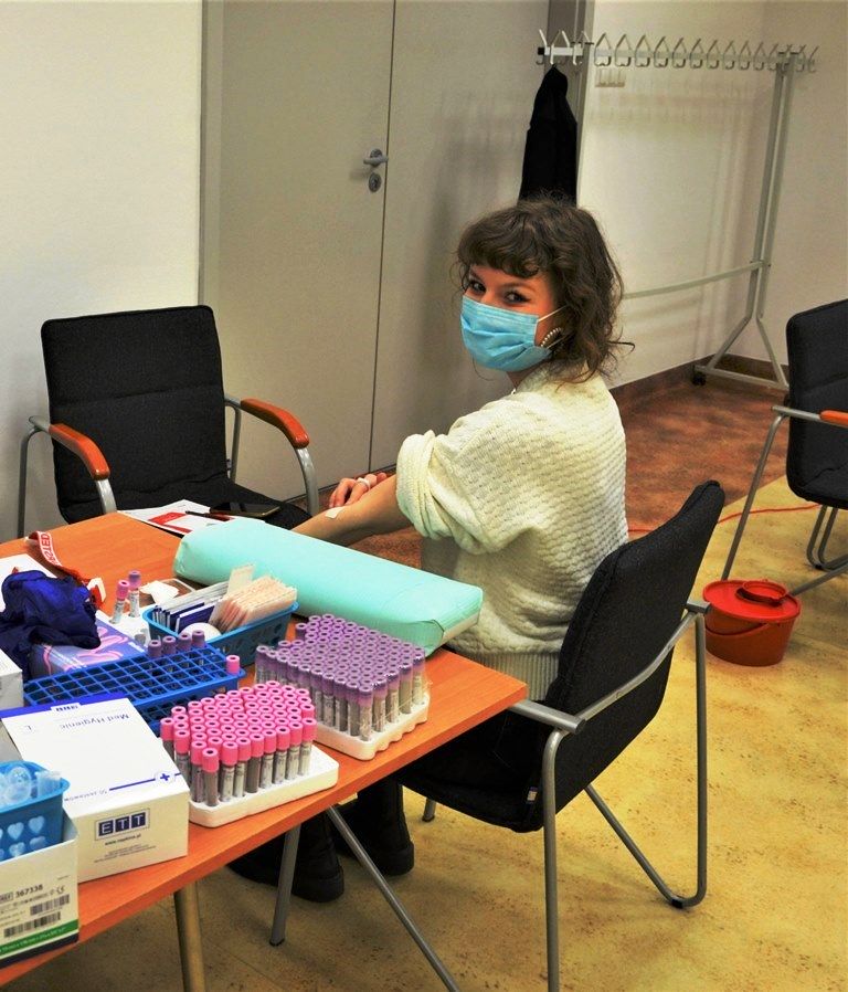 1. kobieta w maseczce siedzi przy stole z probówkami, uciskając miejsce po pobraniu krwi do badania