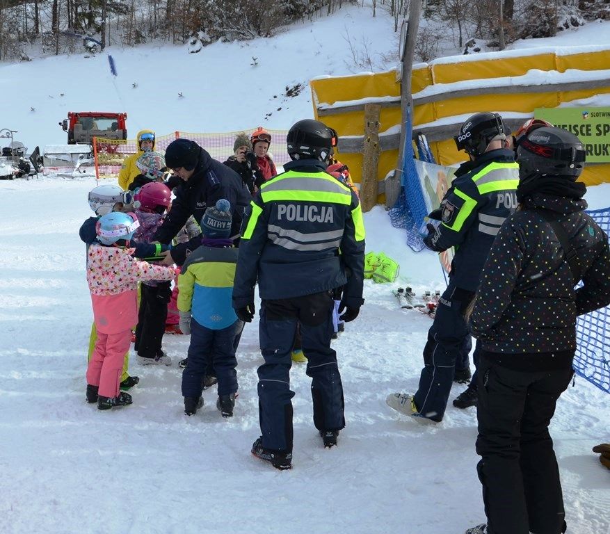 grupa małych dzieci w strojach narciarskich, jednemu z nich policjant zakłada na rękę odblaskową opaskę